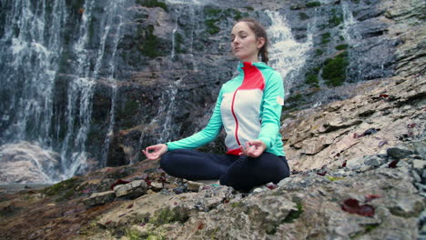 mujer meditando, sentada en posición de yoga de loto junto a una cascada, toma de ángulo bajo