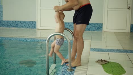 padres enseñando a su hijo a nadar en la piscina. familia joven en la piscina enseñando a su hijo a nadar. padre fuera de la piscina, pone al niño en el agua, madre ayudándole a nadar. en el interior. cámara lenta