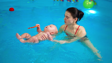 sweet caucasian infant baby lies on the water in the swimming pool. female coach talking to a kid and moving him in water.