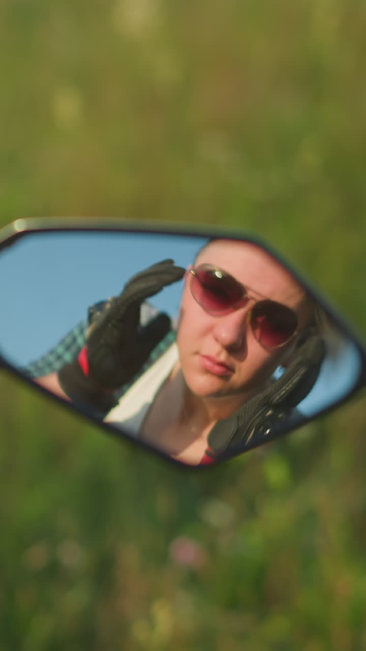 Mirror reflection of a woman adjusting her sunglasses while wearing gloves, with a blurred grassy field in the background, she looks focused, with sunlight softly illuminating her face