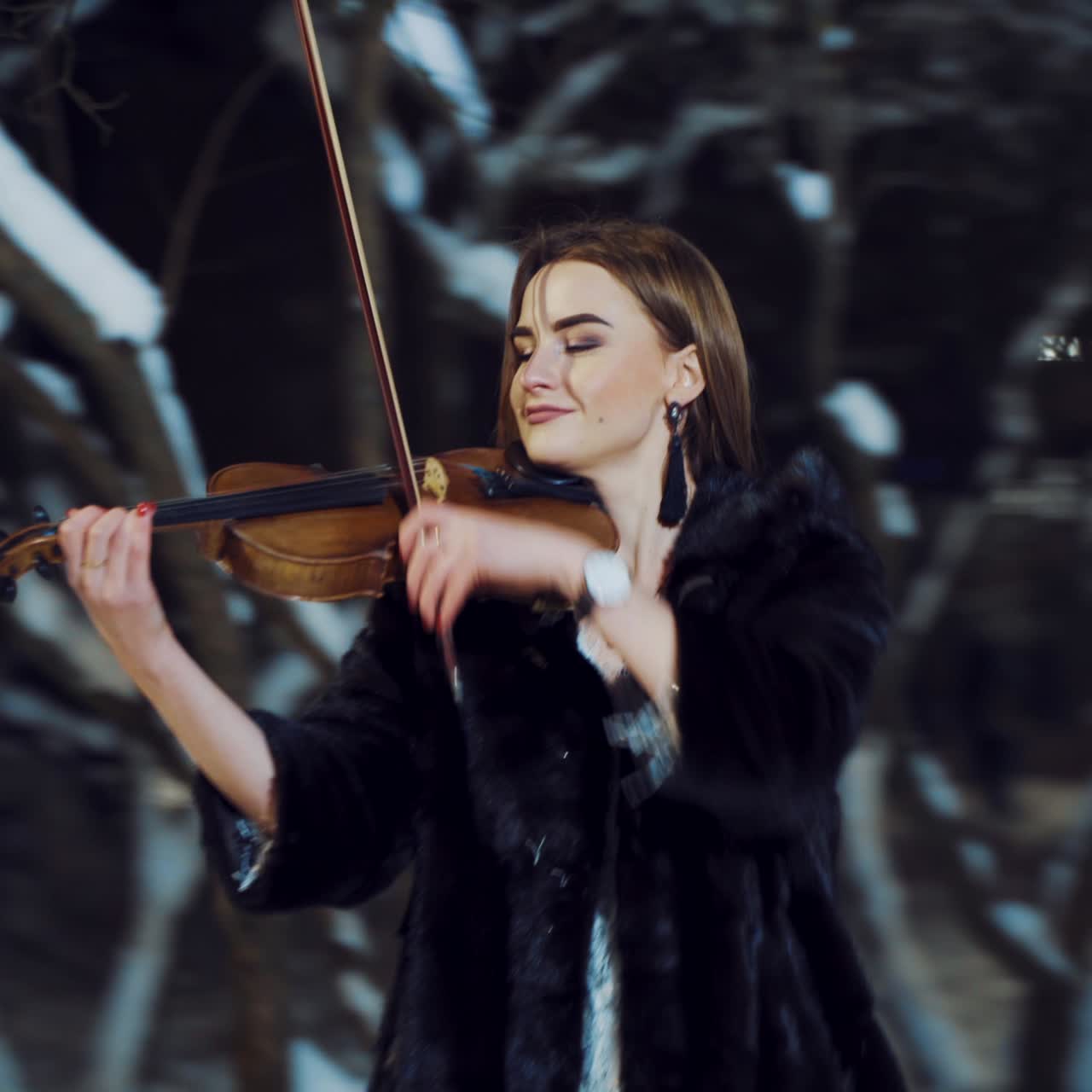 A beautiful girl with make-up and in a white dress is playing a violin composition in a winter park on the background of snow. Camera motion to left