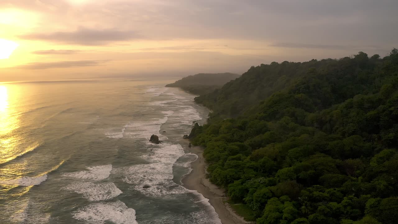 An aerial view captures the tranquil sunset over Santa Teresa Coast in Costa Rica, with waves crashing along a sandy shore framed by lush green hills