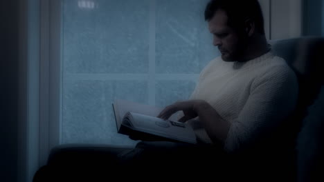 Hombre-Leyendo-Un-Libro-Junto-A-La-Ventana-Por-La-Noche,-Nevando-A-La-Luz-De-La-Luna