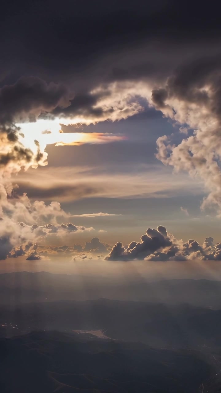 Vertical video: Shifting dark cloud opening over hills from plane as sunlight breaks forming rays