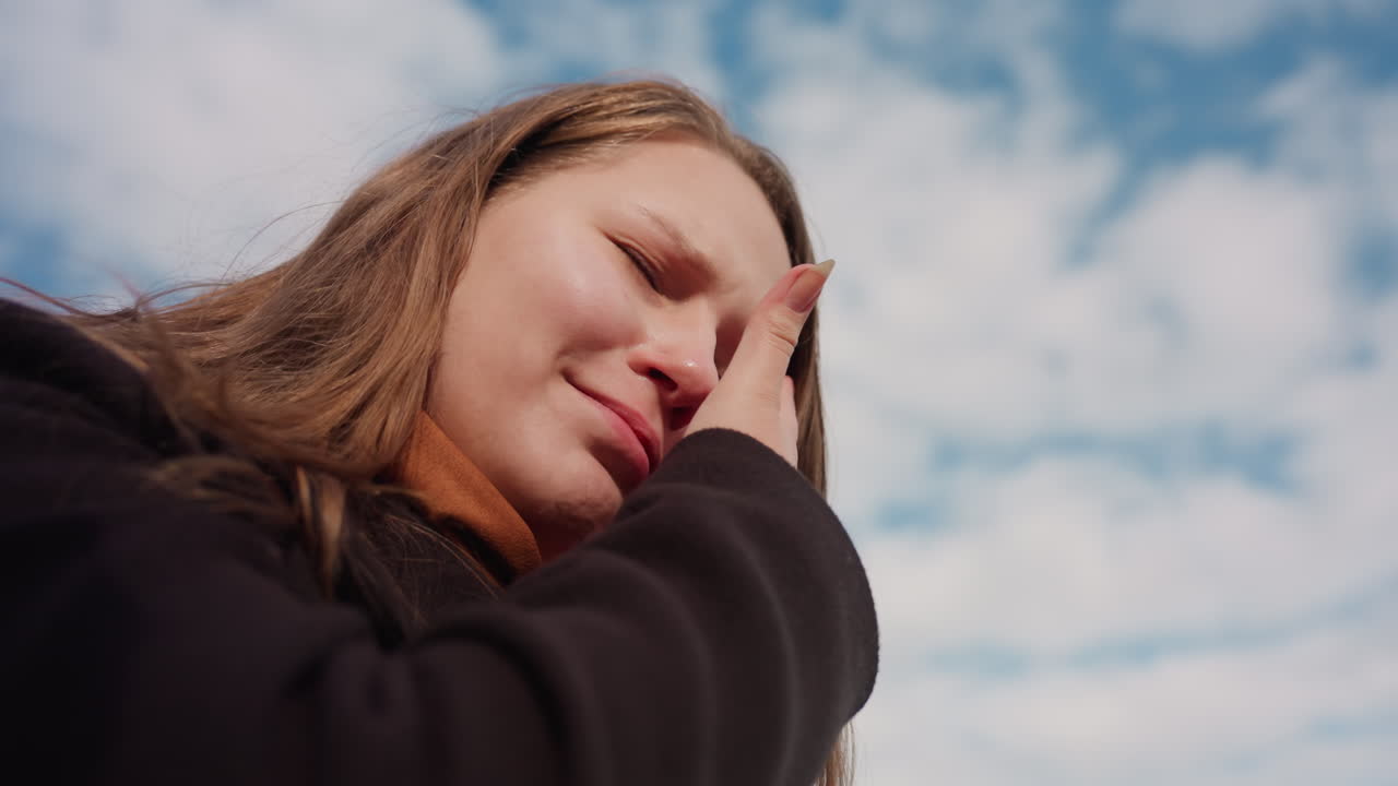 Under view of woman raising eyeglasses toward bright blue sky with soft clouds, sunlight touching skin and hair, creating peaceful atmosphere that highlights natural beauty and sense of open freedom