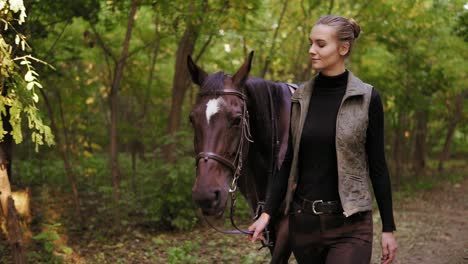 attractive female jockey is petting a stunning brown horse with white spot on forehead while walking together in the forest