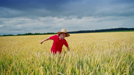 cute boy on yellow field. teenager in red t-shirt and straw hat walking in the golden wheat field under dark sky. summer country life and importance of agriculture.