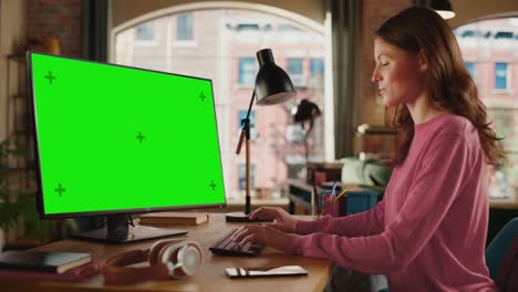 joven mujer adulta hermosa trabajando desde casa en una computadora de escritorio con pantalla verde. mujer creativa comprobando y escribiendo correos electrónicos. apartamento loft con vista a la ciudad urbana desde una gran ventana.