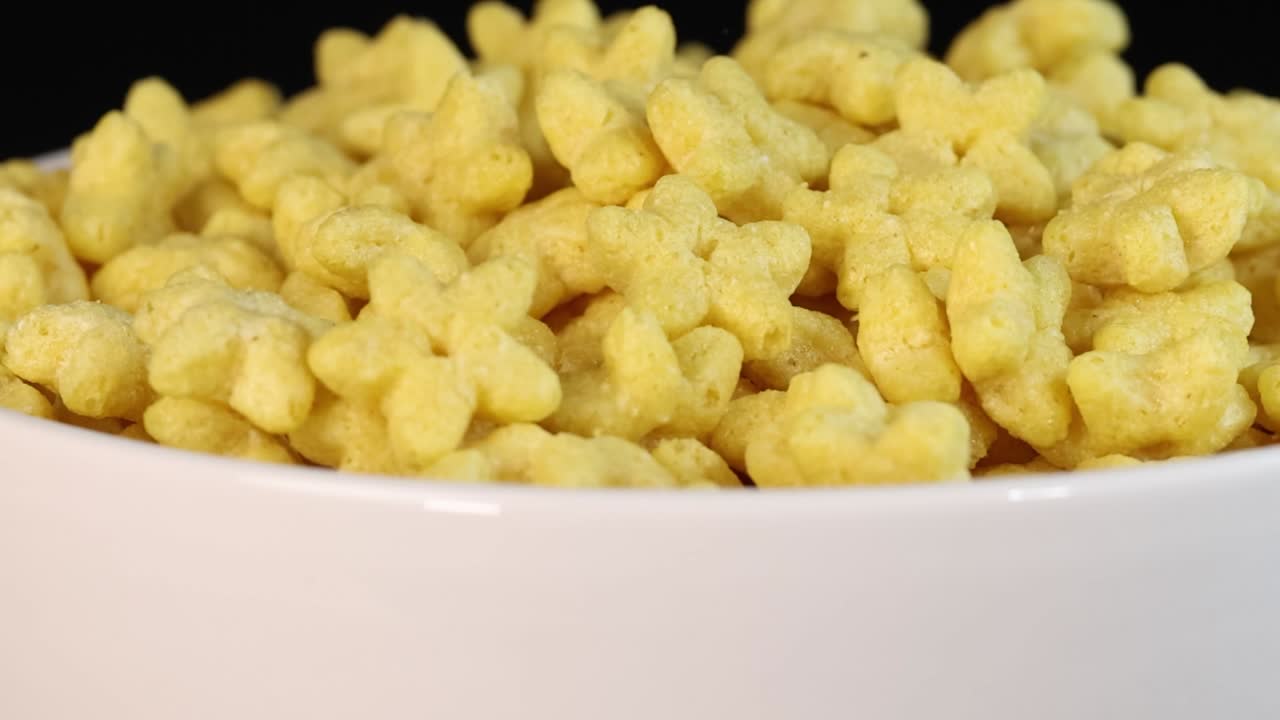 Pezzi di cereali gialli a forma di stella cadono e riempiono una ciotola bianca su uno sfondo nero.