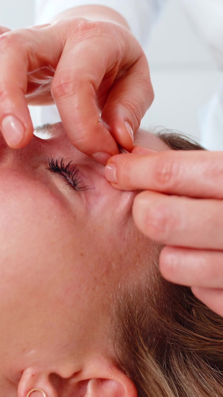 Jovem mulher recebendo massagem facial na área dos olhos por cosmetologista profissional em clínica de estética relaxante