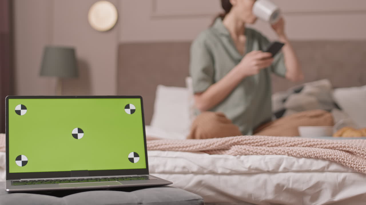 Woman using phone and drinking coffee in bed with laptop
