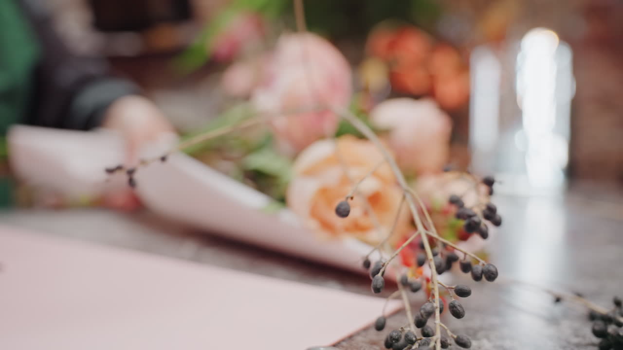 カウンターに置かれたピーチ色のバラの花束のクローズアップ。職人がラッピングペーパーの上でベリーや小枝を使って茎を整えている。柔らかな暖色系の照明が当たるヴィンテージな工房の雰囲気の中で、繊細な花びらや葉が際立つ。