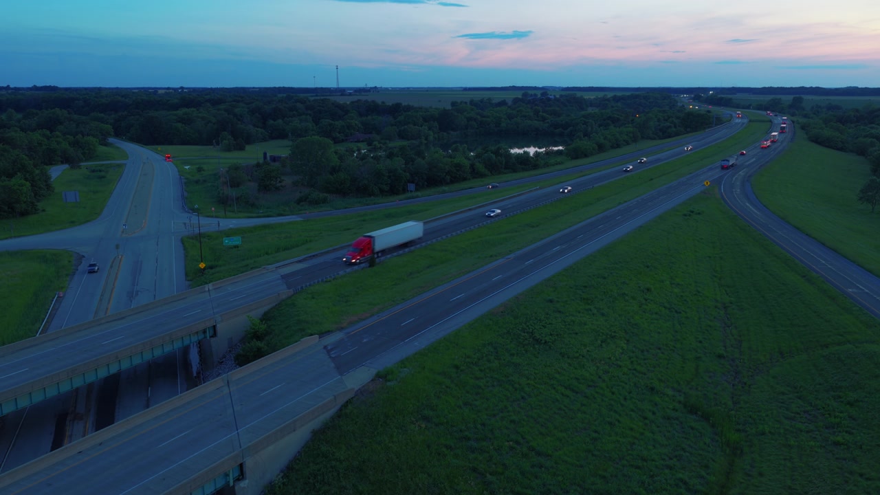 A red semi hauls its trailer across a rural highway bridge as dusk settles — aerial b-roll for trucking, night logistics and delivery-deadline stories.