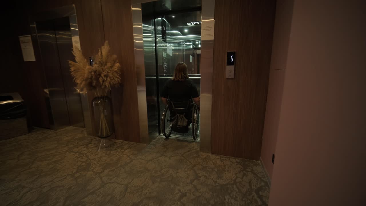 En kvinde i kørestol nærmer sig en elevator i en hotellobby, der viser tilgængelige faciliteter og moderne design. Scenen understreger mobilitet, uafhængighed og inklusion inden for gæstfrihed