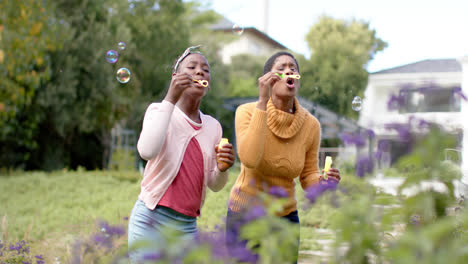 happy african american daughter and mother blowing bubbles in sunny garden