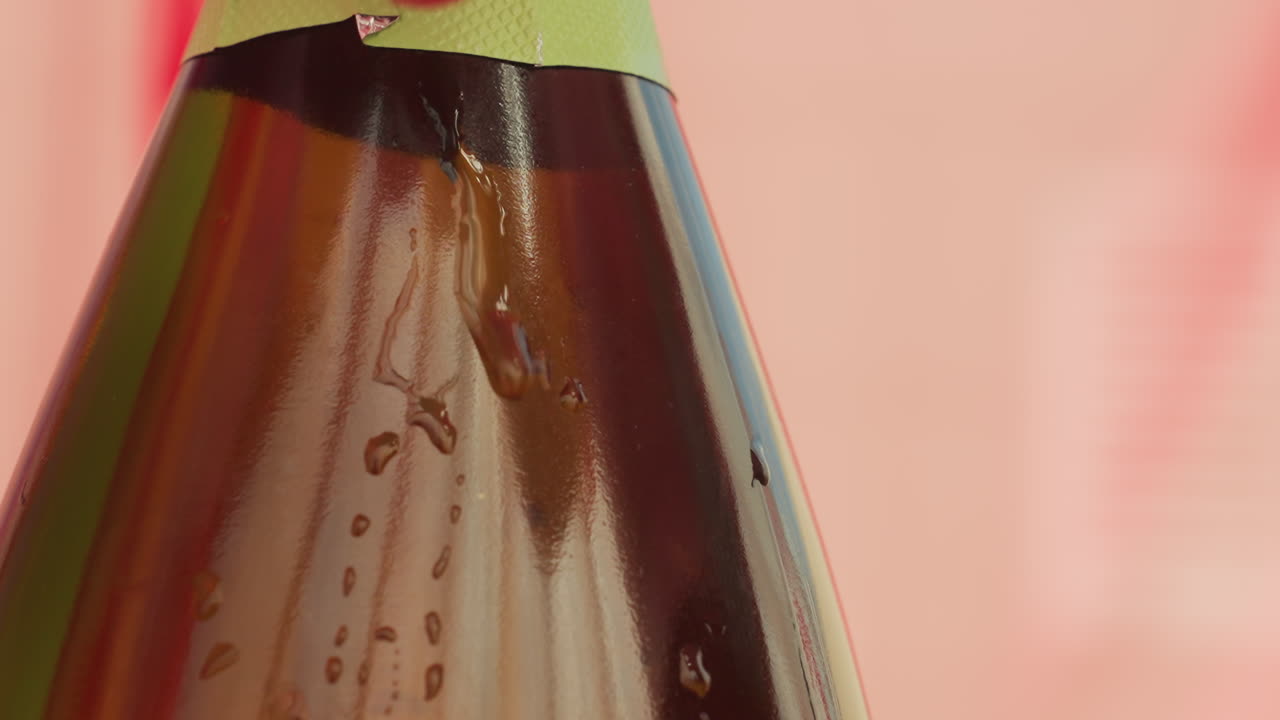 Primo piano di una bottiglia di birra ghiacciata con condensa e tappo dorato, gocce d'acqua su vetro ambrato, sfondo rosa chiaro sfocato, luci da studio, lenta panoramica che suggerisce un ristoro estivo