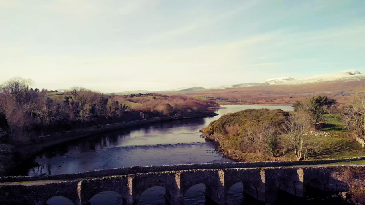 Pullaway from old stone bridge in Mayo, Ireland, with snow capped mountains in the background. Blue sky and water, yellow grass on the banks