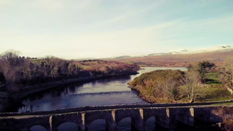 pullaway from old stone bridge in mayo, ireland, with snow capped mountains in the background. blue sky and water, yellow grass on the banks