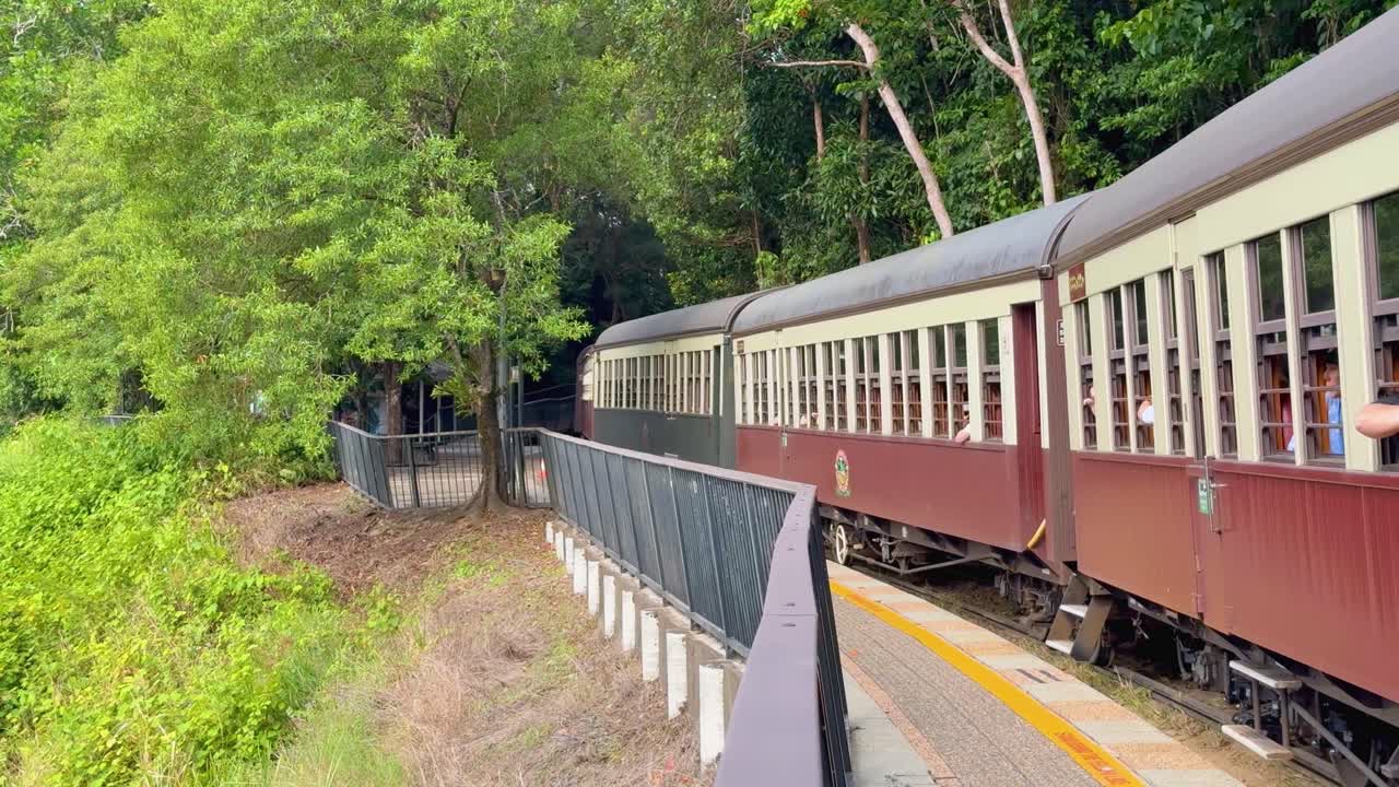 A vintage passenger train travels slowly along a curved track through dense green forest, captured in daylight with steady camera movement and vibrant natural lighting