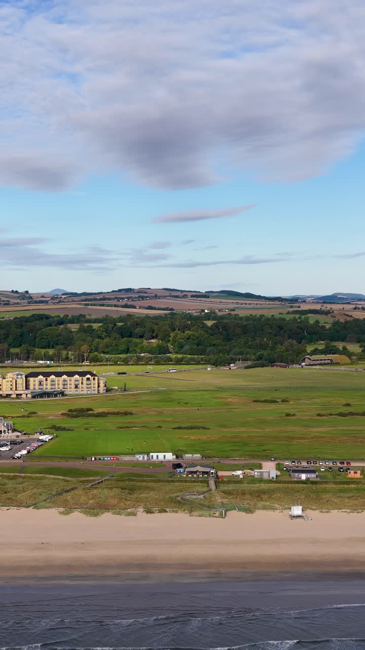 Drone glides above St Andrews, capturing golf course, sandy beach, coastline, and town under daylight