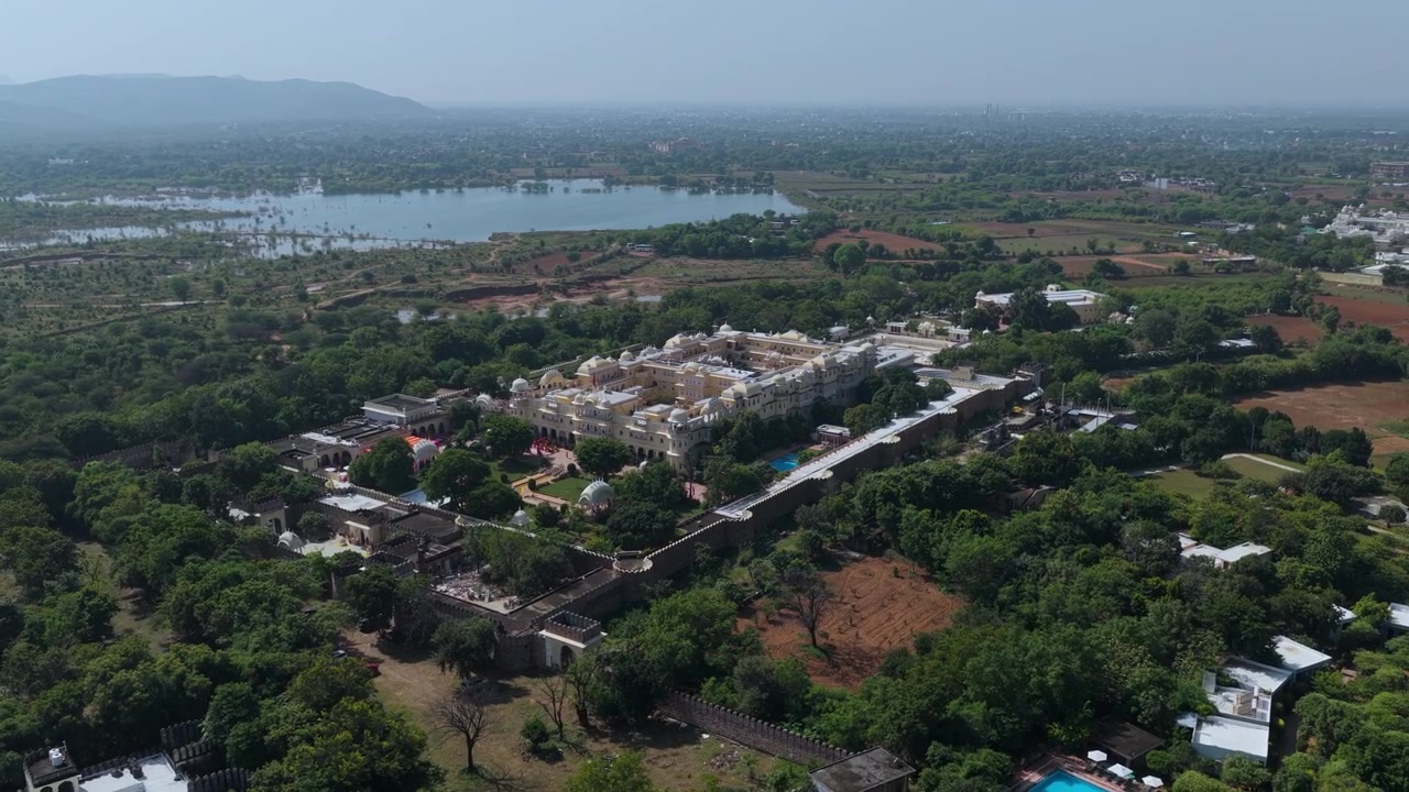 Sebuah bidikan sinematik sudut tinggi yang menampilkan istana warisan putih megah dikelilingi oleh tanaman hijau subur dan danau yang tenang di pedesaan India.