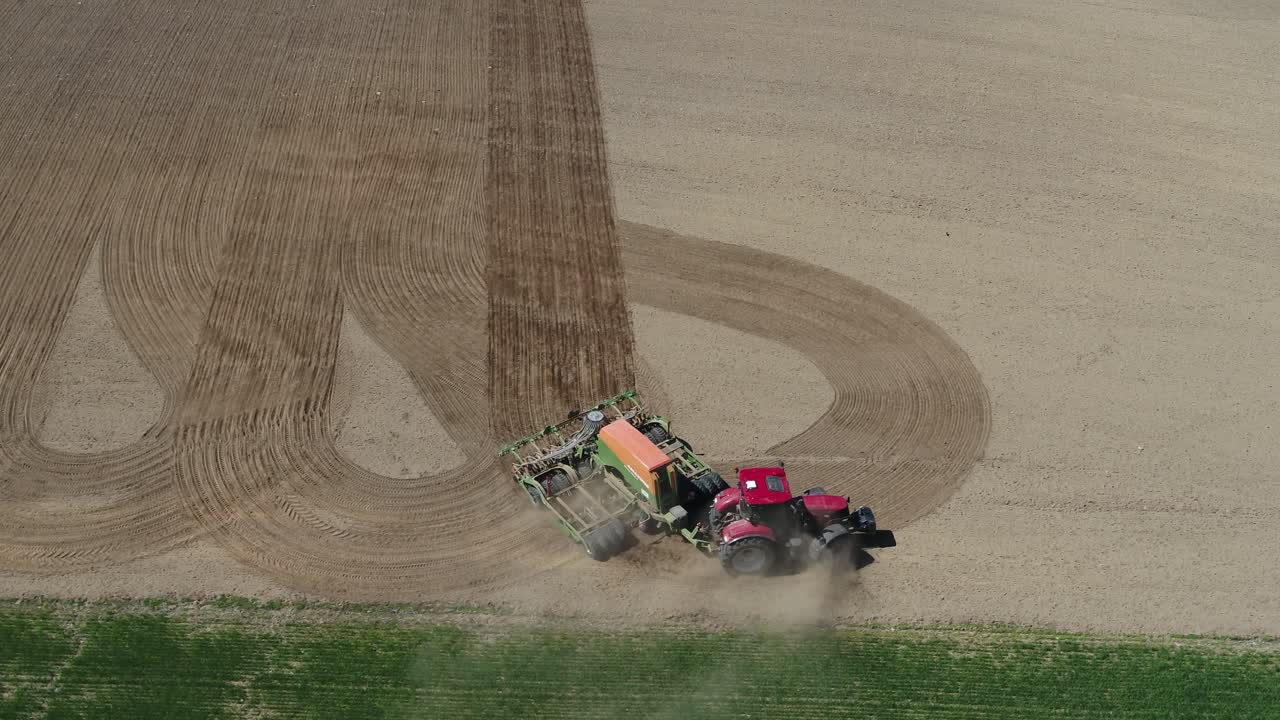 Drone follows red tractor sowing crop lines across brown dirt field under clear blue sky