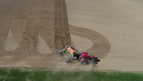 drone follows red tractor sowing crop lines across brown dirt field under clear blue sky