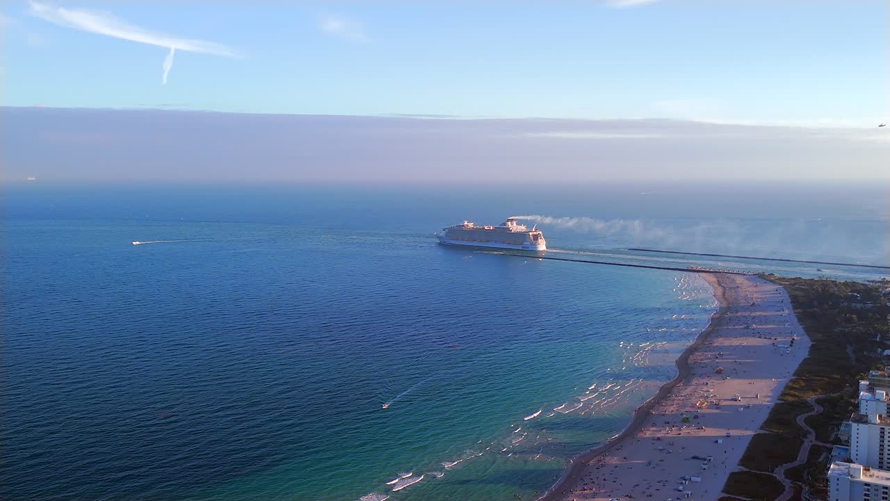 Drone captures a cinematic view of a cruise ship exiting the Port of Miami into the Atlantic Ocean. Flying over the city, it showcases the beach, sand, and the vast horizon from a stunning height.