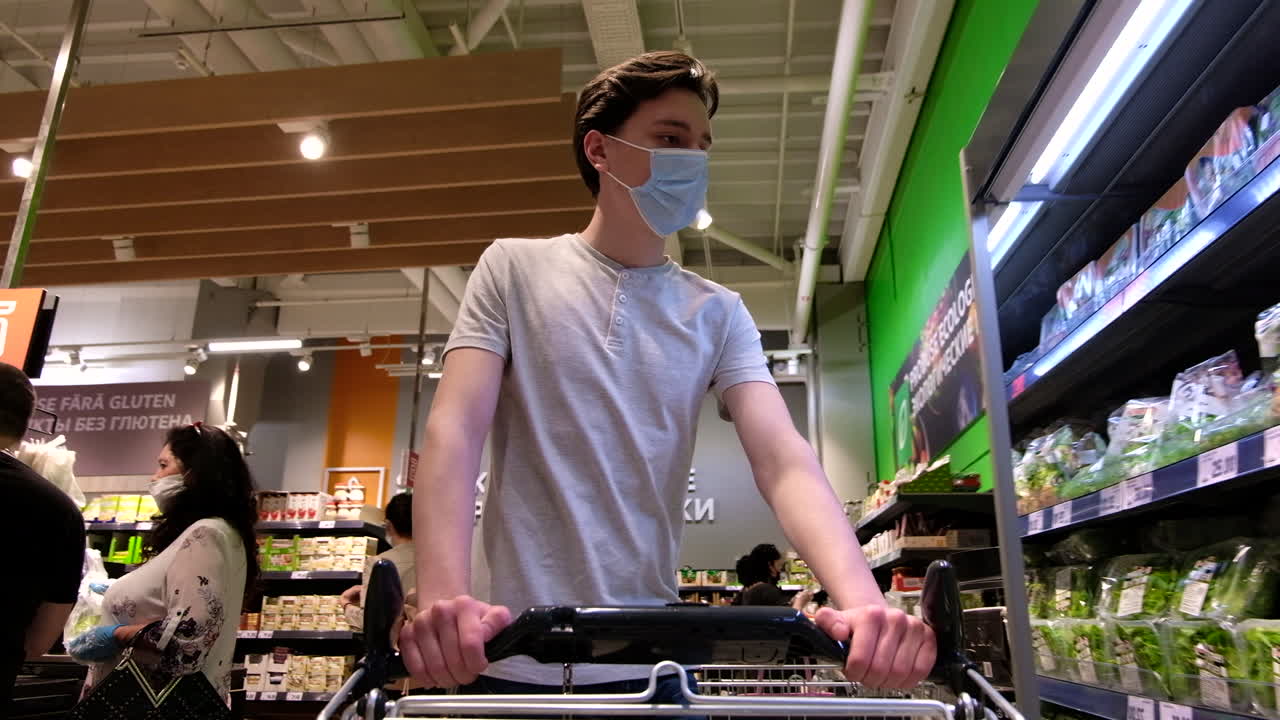 CHISINAU, MOLDOVA - JUNE 5, 2020: Young man with a protective medical mask walking in a supermarket and take greens from the shelf. Corona Virus idea