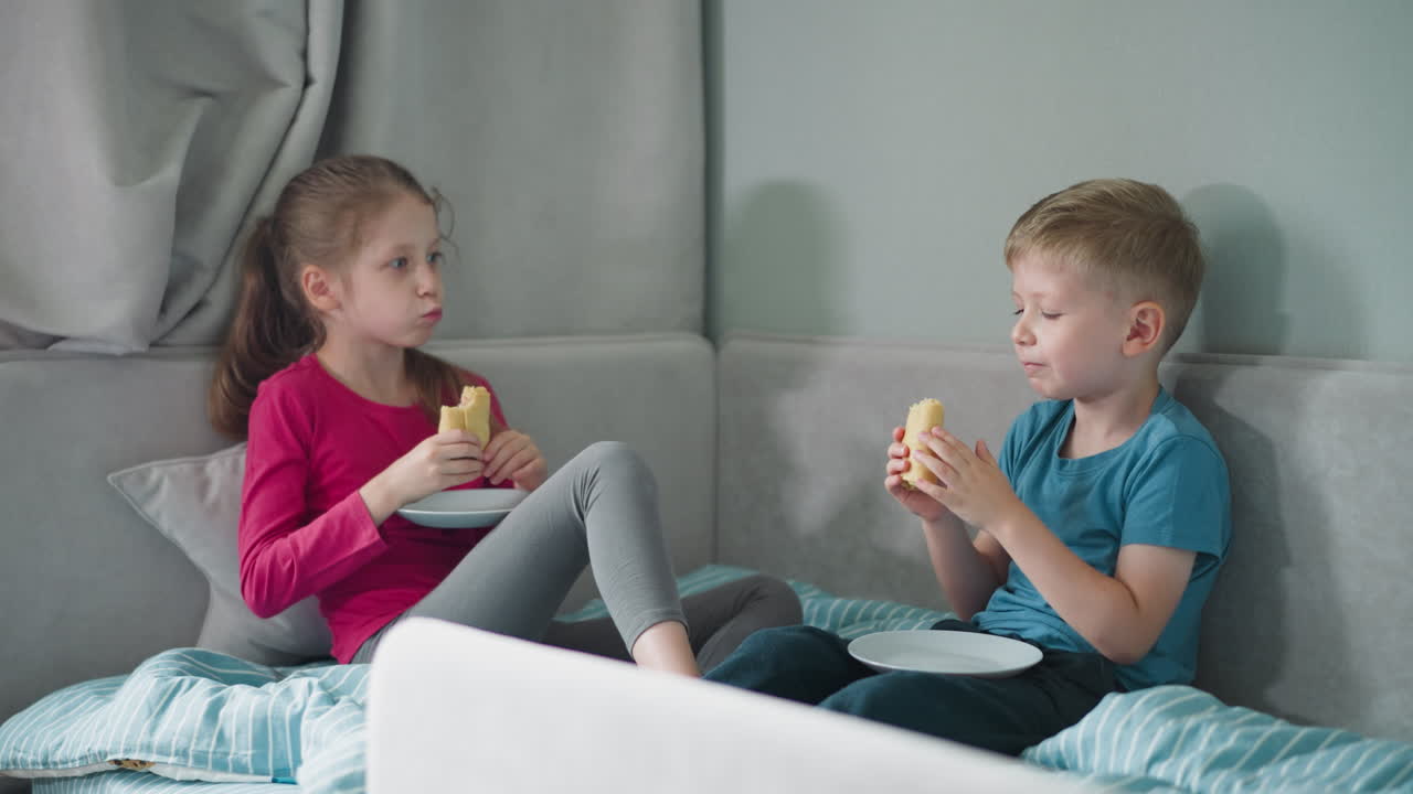 Little boy and girl seated on soft cushioned sofa having conversation while enjoying fish pie together with white plates on laps inside cozy bedroom setting