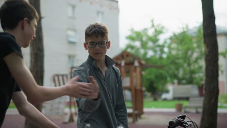 dos hermanos en bicicletas interactúan lúdicamente mientras están en un parque, un hermano en una camiseta gris está sonriendo mientras se dan la mano, en el fondo, un patio de recreo y edificios residenciales