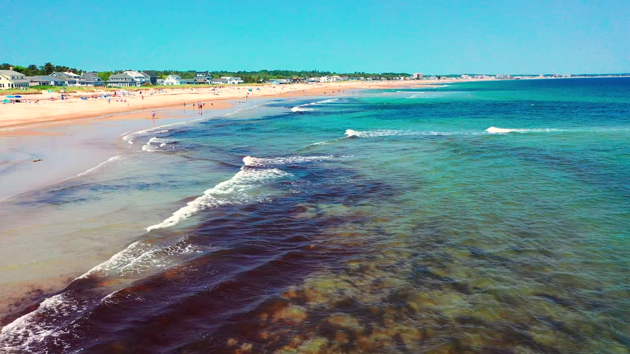Aerial drone video of Saco, Maine coastline in summer. Tourists fill the sandy beach near vacation houses, waves hit shore, families relax. A beautiful East Coast getaway in full peak-season swing