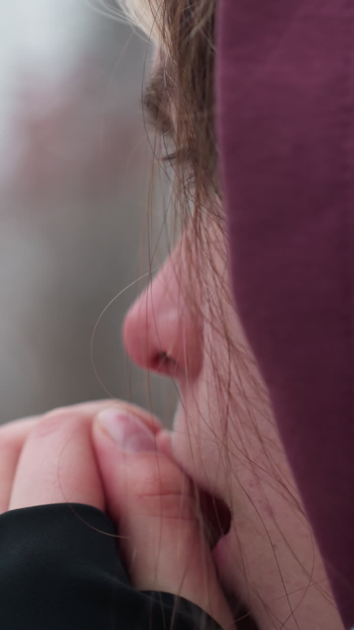 Close-up side face view of young woman rubbing nose with black long sleeve in cold weather, her flushed skin highlights the winter chill, blurred background of trees