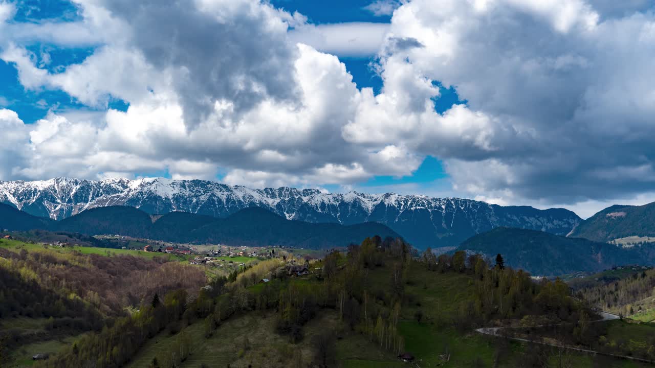 Timelapse af den majestætiske Piatra Craiului bjergkæde med sneklædte toppe, en klar blå himmel med hurtigtsejlende bløde hvide skyer og frodig grøn vegetation, der dækker nærliggende bakker, Rumænien