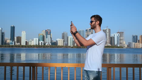 happy handsome man in sunglasses and white t-shirt with a beard taking photos on a smartphone while standing on the waterfront in the summer town in the background and buildings