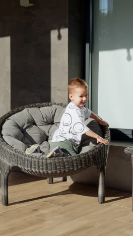 little baby boy comes up to the comfortable armchair standing outdoors at the porch. kid climbs into the chair and sits down cozily. vertical video.