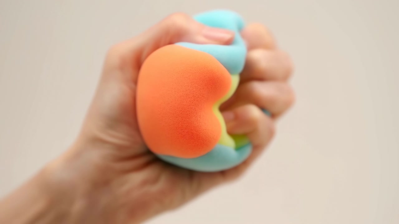 Close view of hand compressing colorful squishy foam toy, revealing soft rounded form with orange, yellow, cyan, and blue sections, neutral beige backdrop, gentle light, calm commercial feel