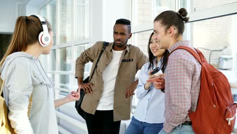 beautiful attractive female student wearing headphones is going down corridor meeting her groupmates giving five to her friend and moving on. she show middle finger to african american guy