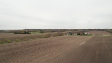 Crows-scatter-and-fly-away-as-drone-approaches-flying-over-newly-seeded-farm-field-with-barn-and-silos-in-distance