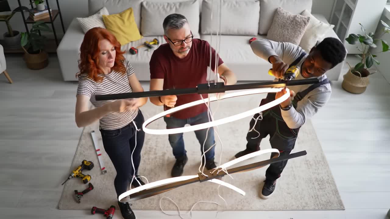 Two individuals are working together to install a contemporary light fixture in a bright living room. They are using tools and coordinating their efforts to secure the setup properly.
