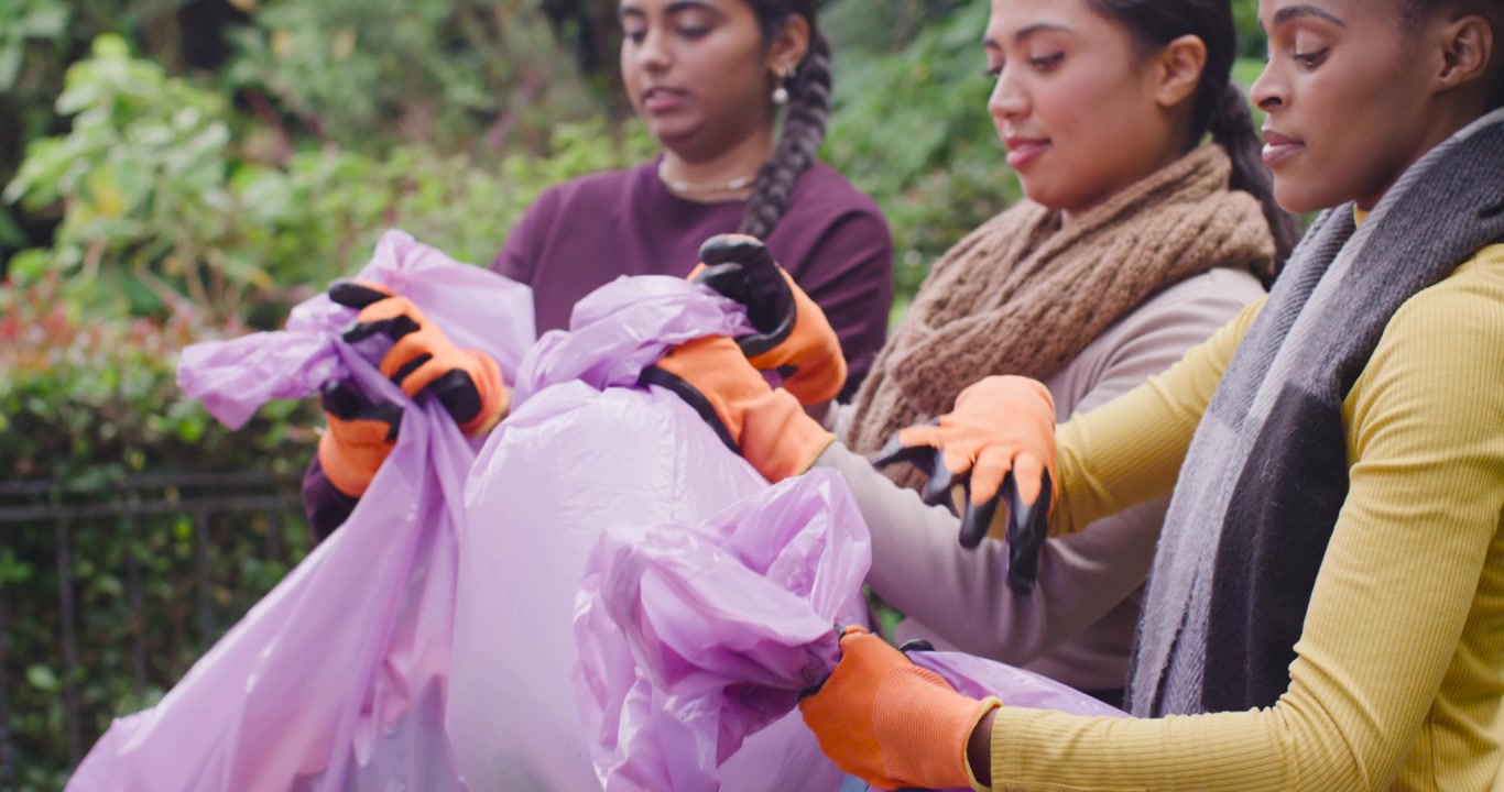 Three women tying lavender trash bags after park cleanup, using orange gloves to seal for pickup