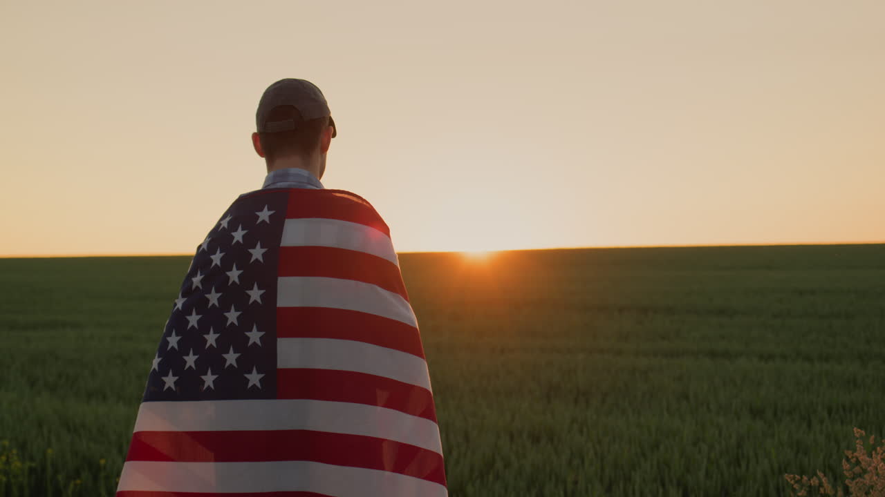 joven granjero con la bandera de estados unidos en los hombros mira el amanecer sobre el campo de trigo