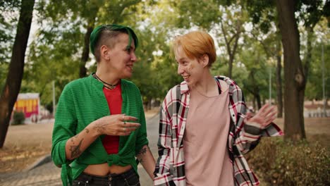 front view of two lesbian girls with bright short hair in shirts walk through the park during their date and communicate