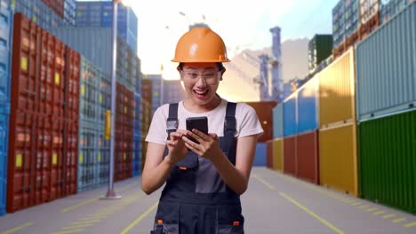 trabajadora asiática con gafas de protección y casco de seguridad mirando su teléfono inteligente y luego gritando celebrando la meta mientras estaba de pie en el almacén del patio de contenedores