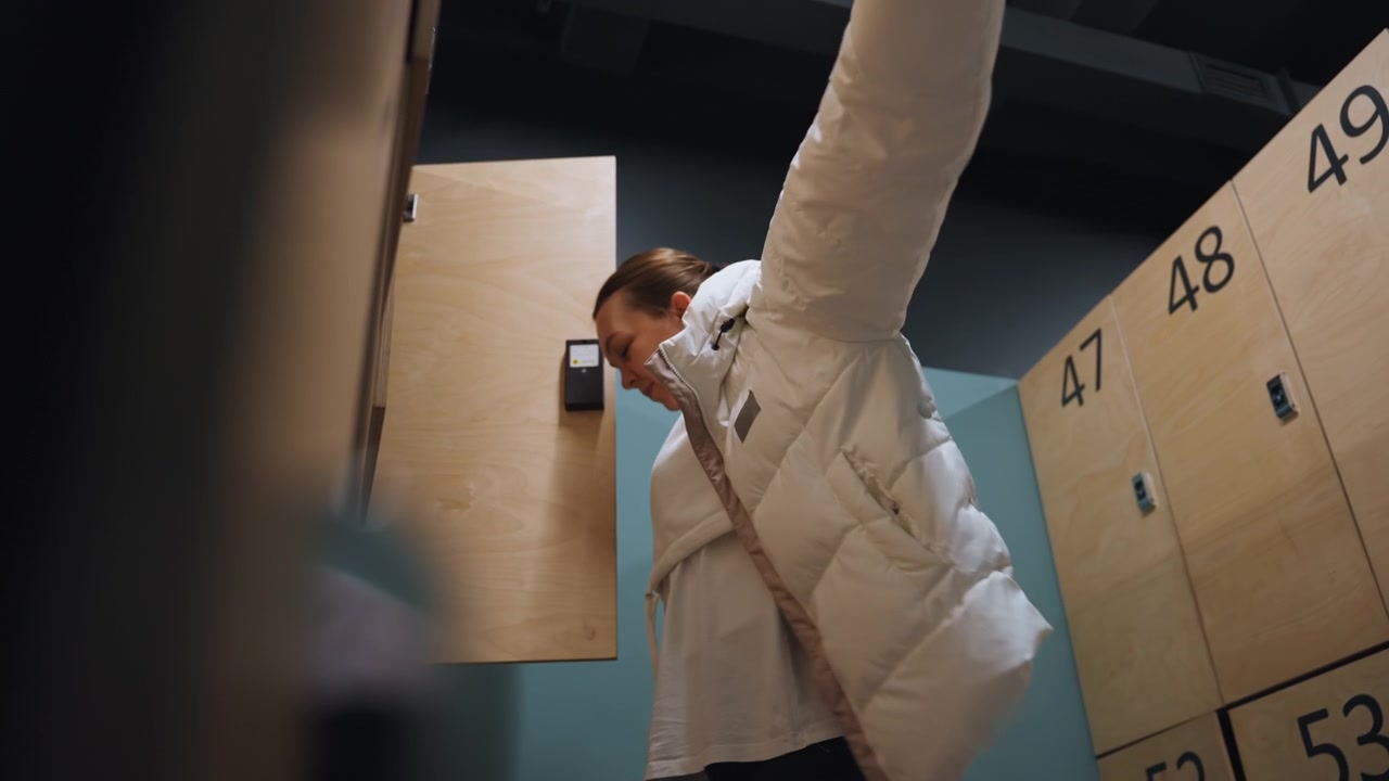 Locker badminton player putting on coat in numbered wooden locker bay, white puffer jacket, ponytail, moody overhead lighting, fastening zipper, checking key and preparing for departure
