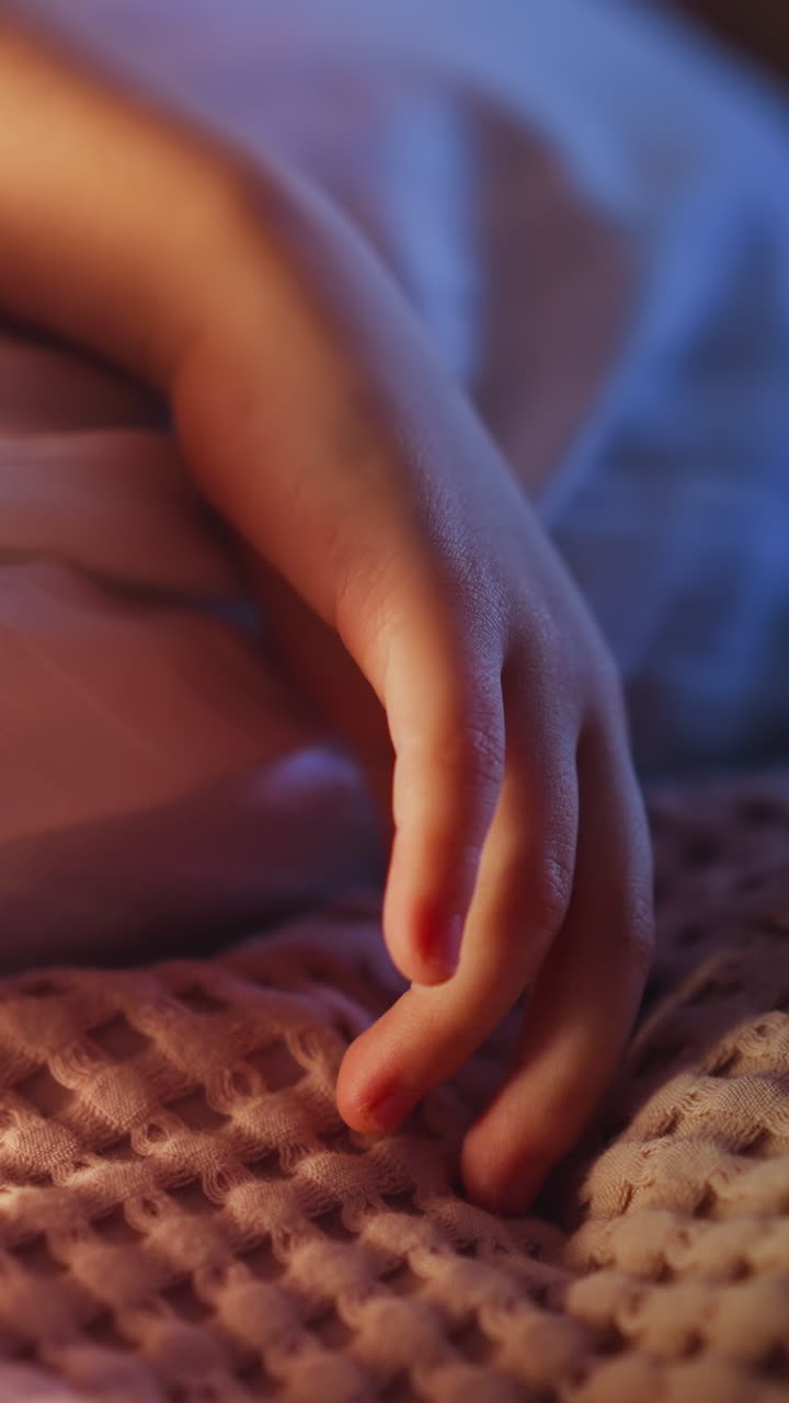 Hand of little child lying on pillow in white case touches pink woolen plaid on bed in semidark bedroom at night extreme close view slow motion