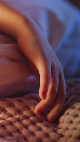 Hand of little child lying on pillow in white case touches pink woolen plaid on bed in semidark bedroom at night extreme close view slow motion