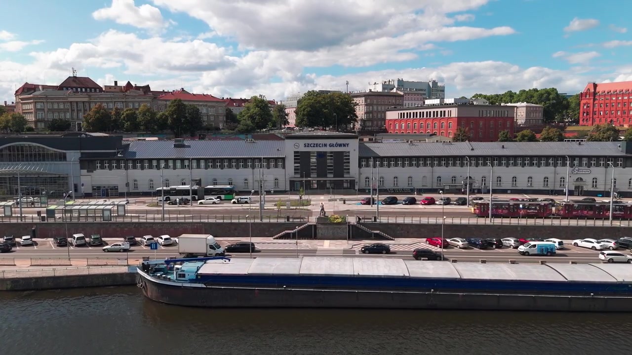 Un'ampia inquadratura della facciata della stazione ferroviaria centrale di Stettino Główny in Polonia, con il lungofiume, una chiatta ormeggiata e il traffico cittadino sotto un cielo blu.