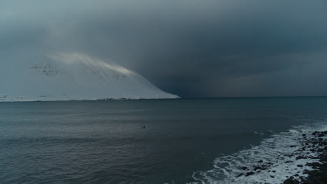 Ein-Surfer,-Der-Im-Winter-Unter-Schneebedeckten-Bergen-In-Island-Sitzt-Und-Mit-Einer-Drohne-Aufgenommen-Wurde