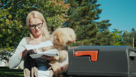 a young woman with a small dog in her arms picks up letters from a mailbox